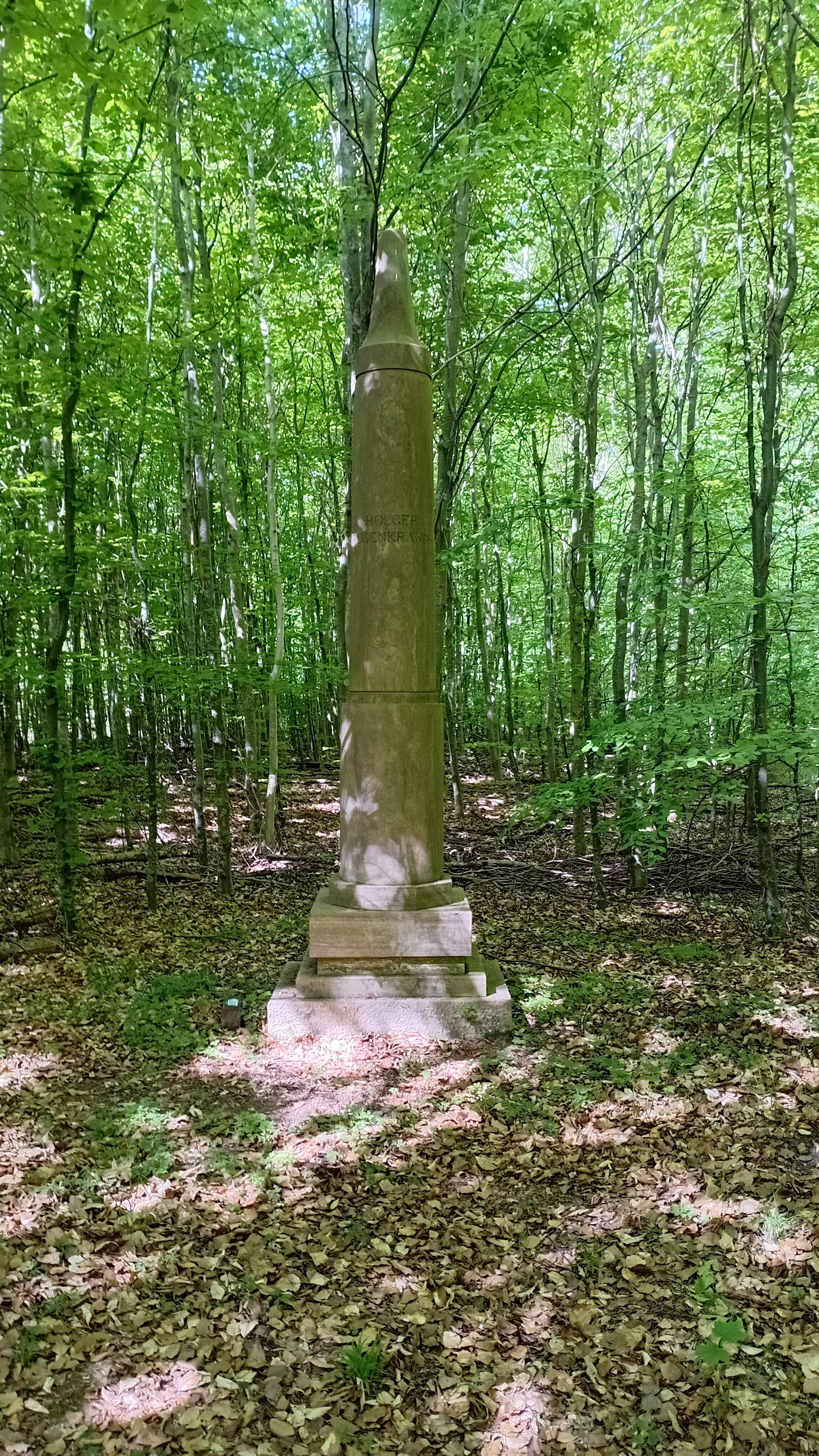 Johannes Wiedewelt: Stele i Mindelunden i Jægerspris. Det over 4 meter høje slanke søjleformede monument for Holger Rosenkrands står blandt skovens træer, og har taget farve efter dem. Foto: Ole Bak Jakobsen.